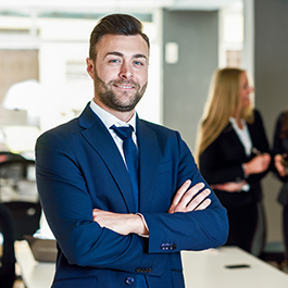 Caucasian businessman leader looking at camera in modern office with multi-ethnic businesspeople working at the background. Teamwork concept. Young man with beard wearing blue suit.